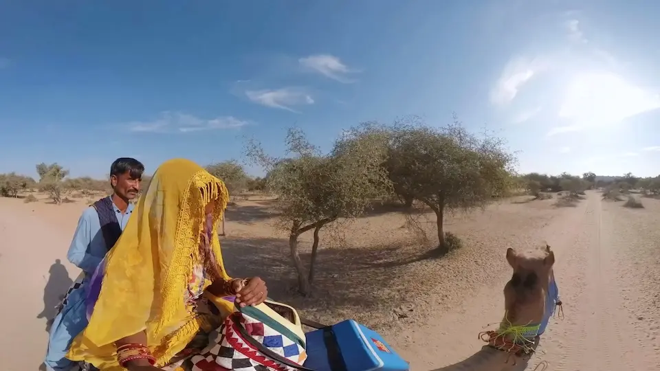 A woman in a yellow head covering holds a small blue cooler while seated in front of a man on top of a camel.
