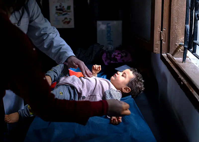 Two people attend a baby who's lit by sunshine coming through a wood frame window.