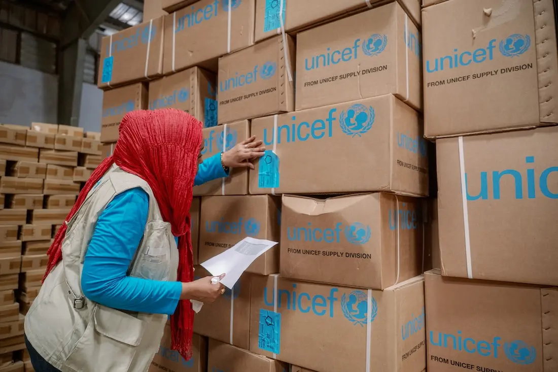 Woman wearing head scarf and vest examines cardboard boxes labeled UNICEF