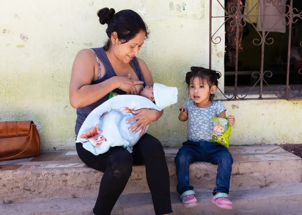 A mother in Guatemala breastfeeds her infant child sitting next to her 3-year-old daughter.