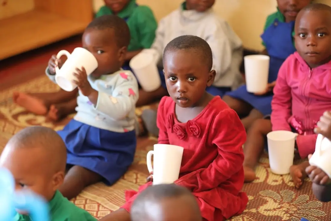 Young girl sitting on floor holding white mug looking directly into camera, surrounded by additional children holding and drinking from mugs