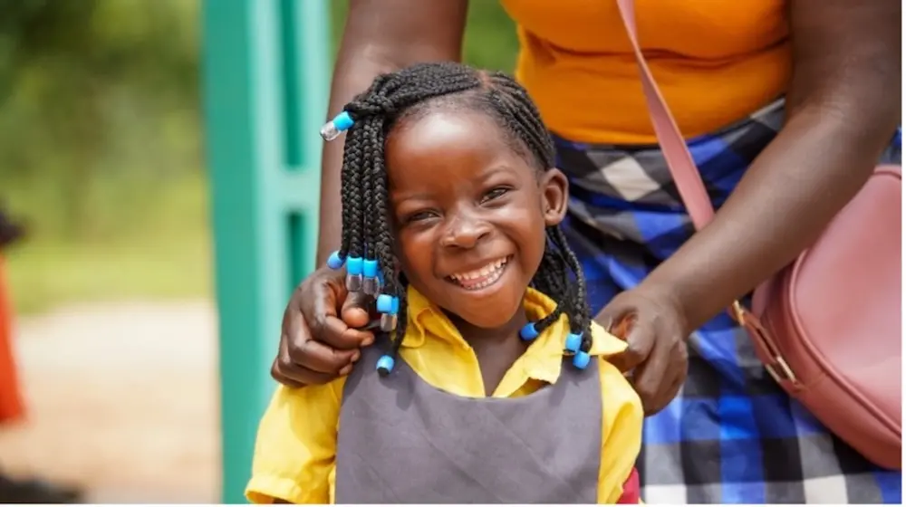 Young girl in school uniform smiles at camera while woman standing behind her placing arms on her shoulder