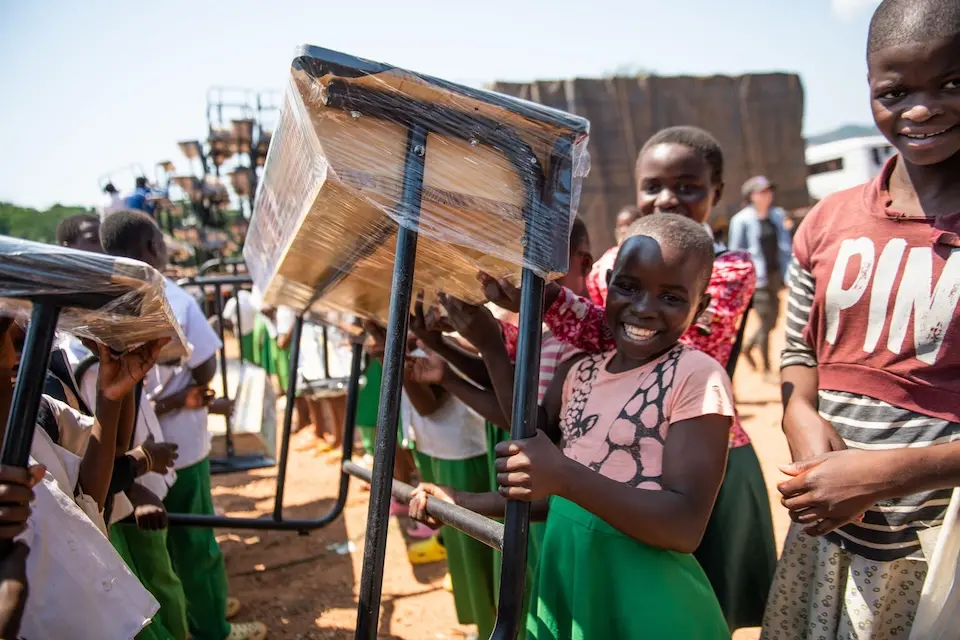 Smiling schoolchildren carry new desks from the K.I.N.D. Fund into Vizara Primary School in northern Malawi in 2024.