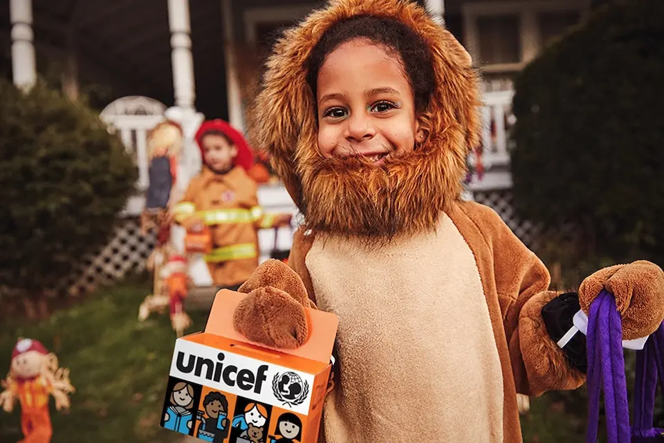 Kid in lion costume holds orange box for Trick-or-Treat for UNICEF fundraising.