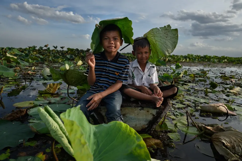 In Thailand during a heat wave in May 2025, two children sit on wet ground holding lotus leaves