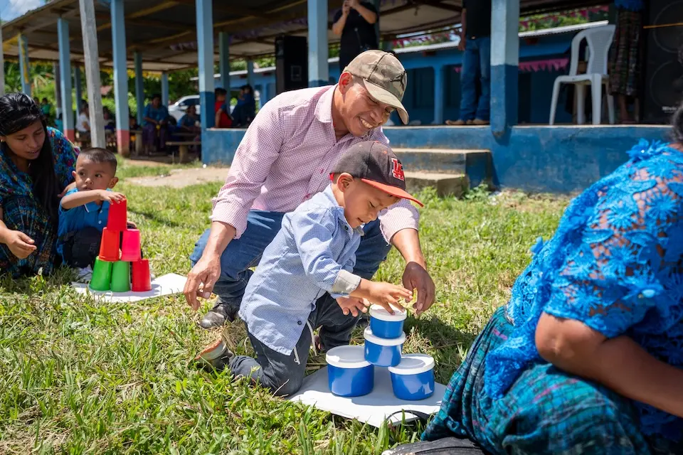 Young boy kneeling and stacking plastic cups while his father kneels behind him and assists