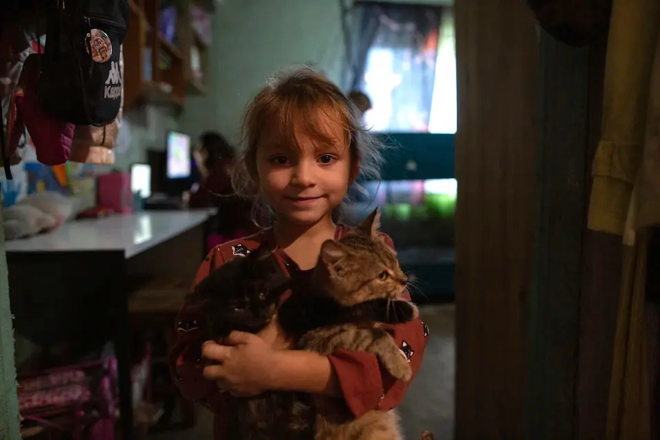 A little girl holds a cat in the dimly lit house in Staryi Saltiv, Kharkiv region, Ukraine. 