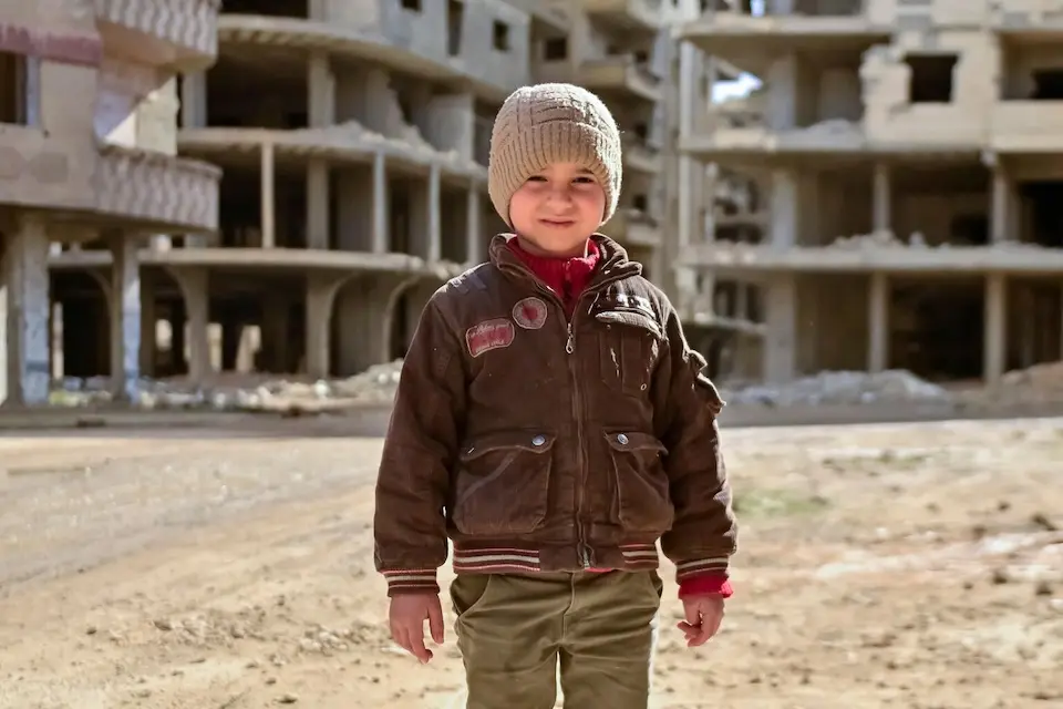 A 5-year-old boy wearing a hat and jacket stands in front of bombed-out buildings in Tadamon, Damascus, Syria on Jan. 14, 2026.