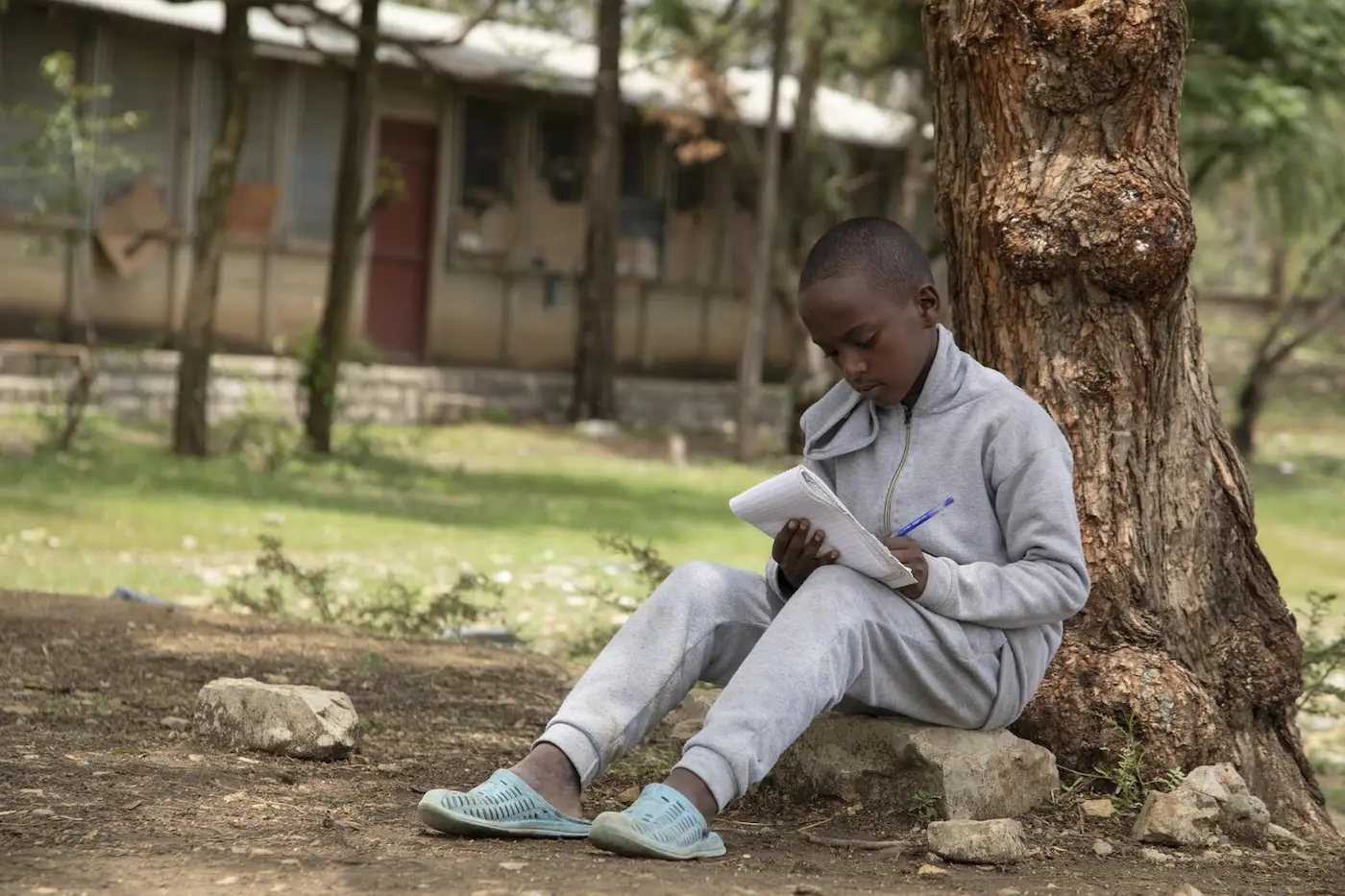 A boy in Ethiopia writes poetry in a notebook sitting by a tree. He uses poetry to help him understand his experiences and feelings.