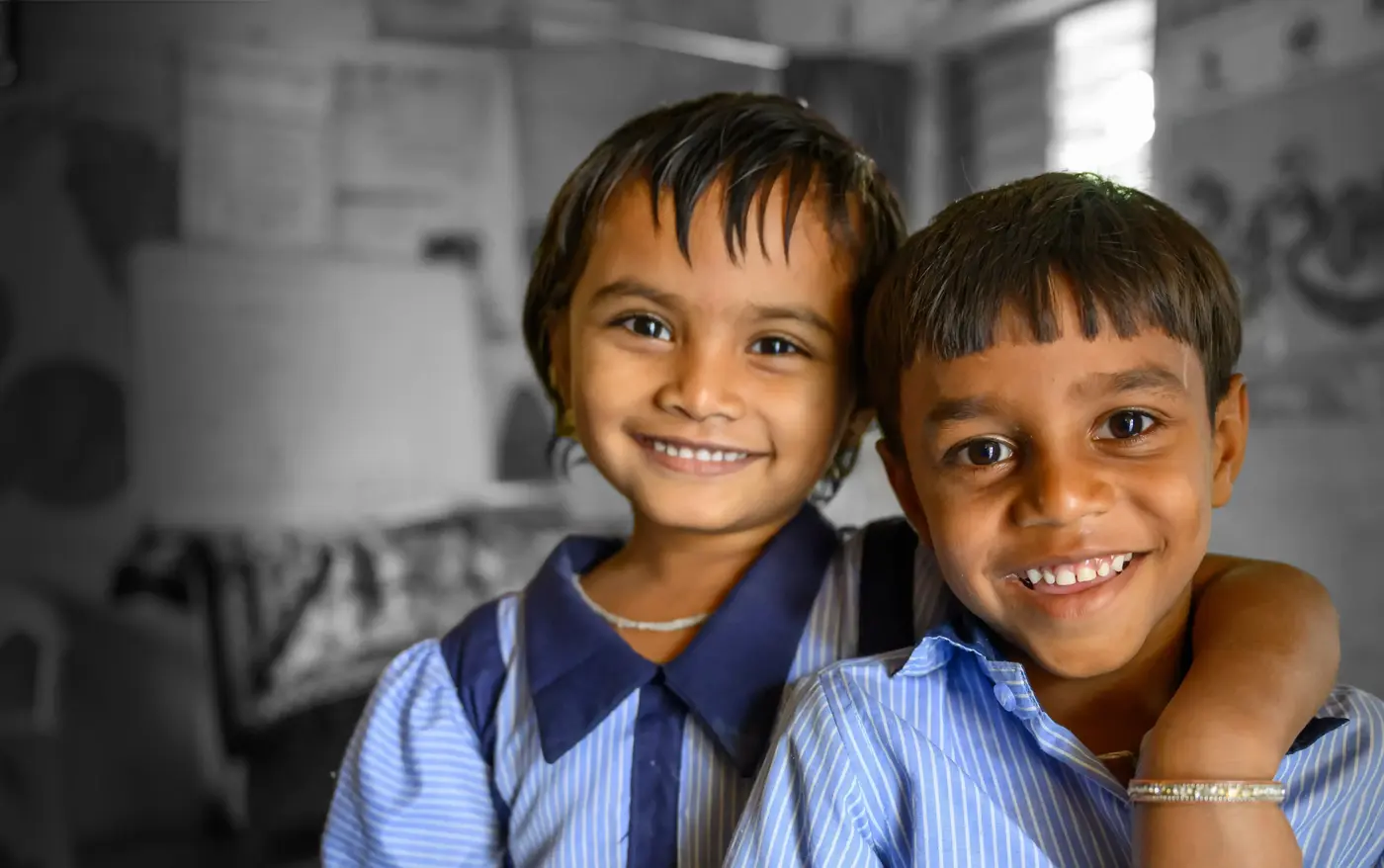 Two children in blue school uniforms smile in a classroom, the girl has her arm around the boy in a friendly way