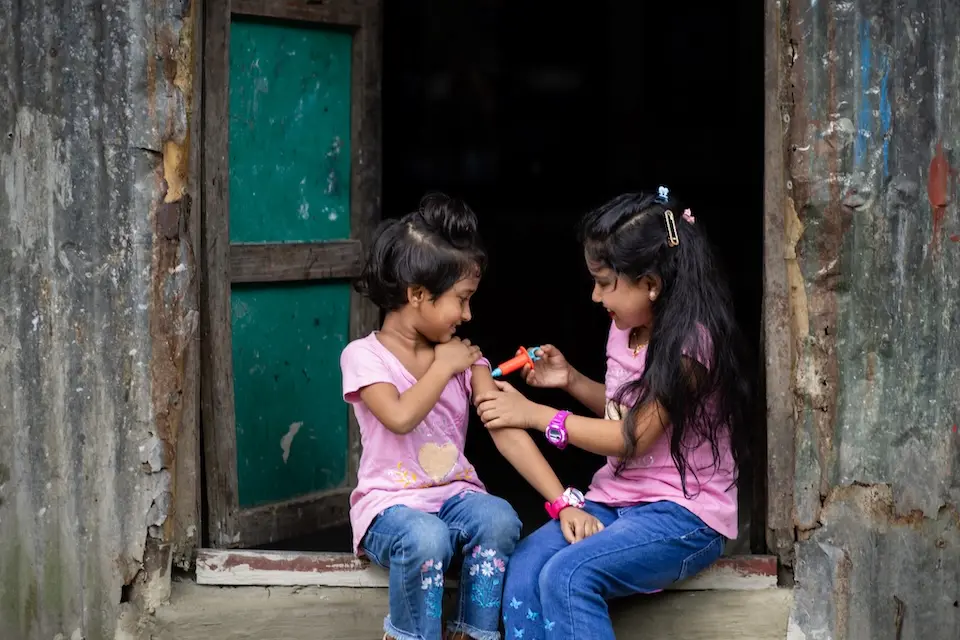 Two girls sitting in a doorway, girl on right holding a plastic toy syringe