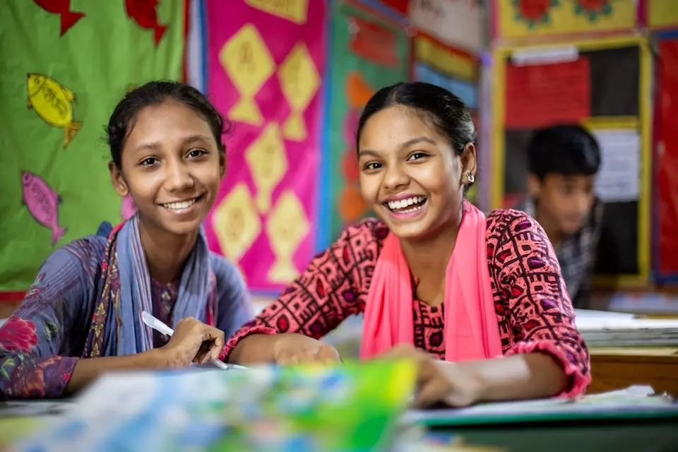 Two girls looking directly at camera and smiling, sitting at table covered with schoolbooks