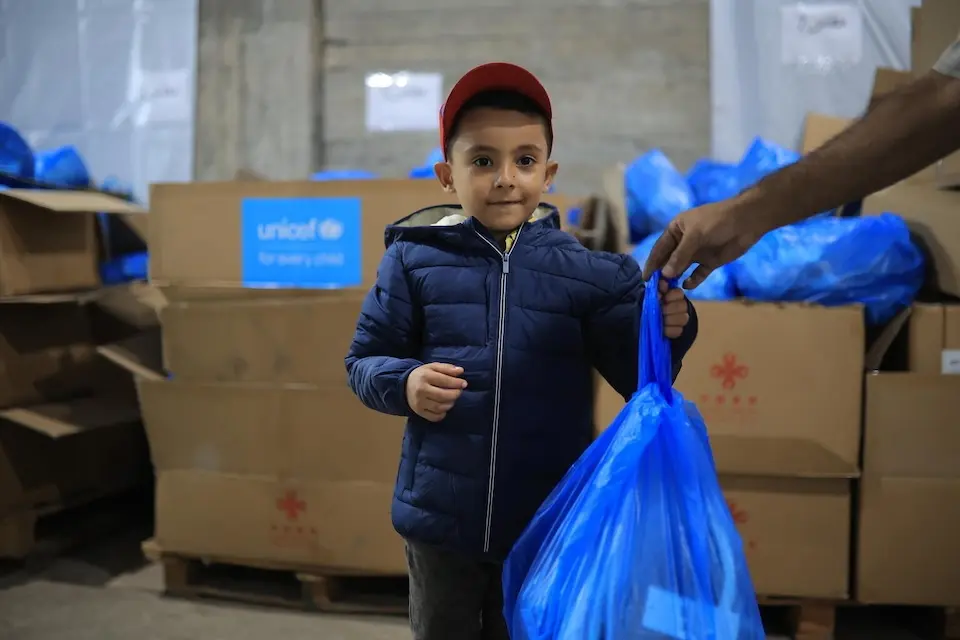 On Oct. 6, 2026 in Gaza, a child wears a warm jacket he received from UNICEF as a staff member hands him a bag of winter clothing.