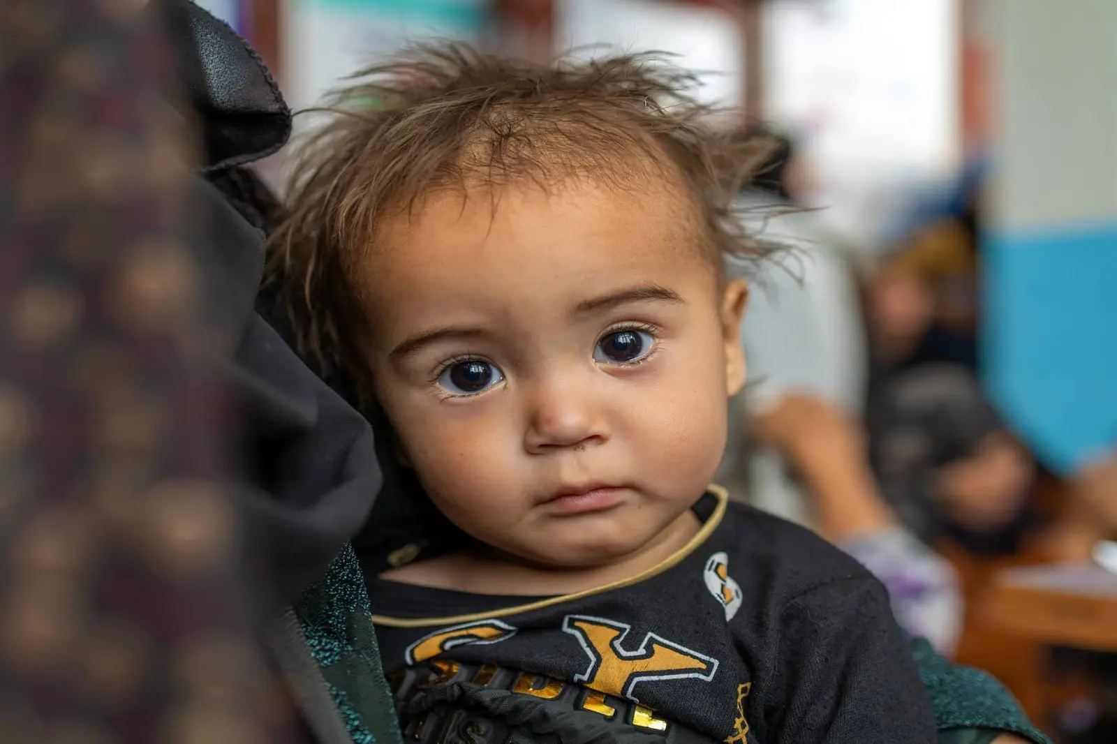 Aref sits on his mother's lap and looks toward the camera while waiting for his nutrition assessment.