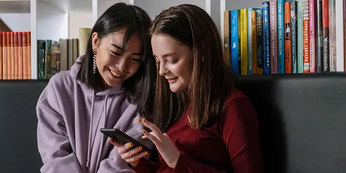 Two girls sitting in front of bookshelves smile looking at a phone together