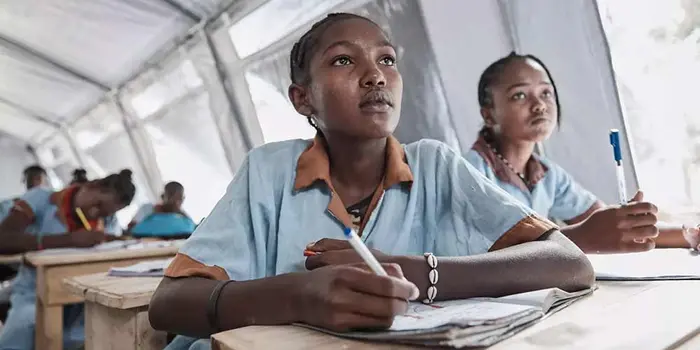 Two girls sit at their desk in front of other students at desks, in a large tent classroom, with their notebooks open, paying attention to a speaker offscreen.