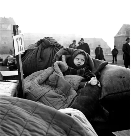 Black and white photo of unattended baby looking wary, bundled in winter clothing and on the ground next to piles of belongings and a sign reading '12', among adults milling about a city square.