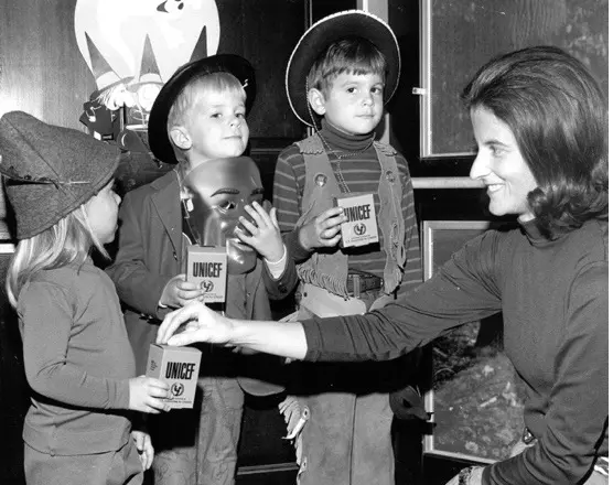 Black and white photo of woman distributing coins to children in costumes holding Trick-or-Treat for UNICEF coin collection boxes.