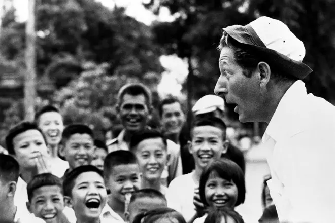 A group of children and adults lauhging and smiling at Danny Kaye, who performs in front of them wearing a tiny hat, making a funny face for the crowd.