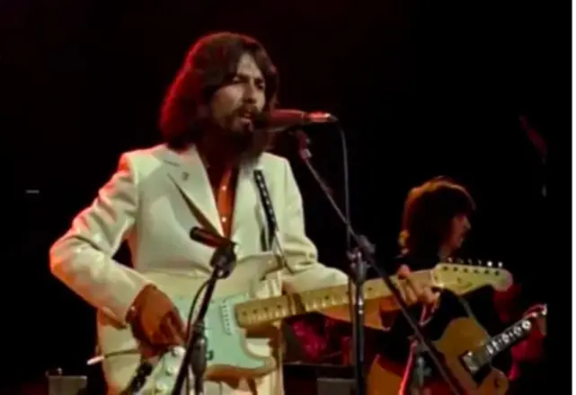 George Harrison, with shoulder-length hair and bushy beard, in a white suit playing a white guitar and singing in front of other musicians on stage.