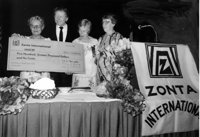 Three women stand with Danny Kaye behind a large banner reading ‘Zonta International’ and hold an oversized $516,000 check from Zonta to UNICEF. 