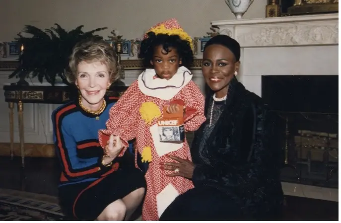 Nancy Reagan and Cicely Tyson kneel on either side of a child wearing a clown costume and holding a Trick-or-Treat for UNICEF collection box.
