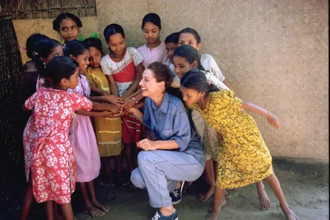 A group of smiling young girls standing in a circle stack their hands in the center. They are joined by a laughing Audrey Hepburn. 