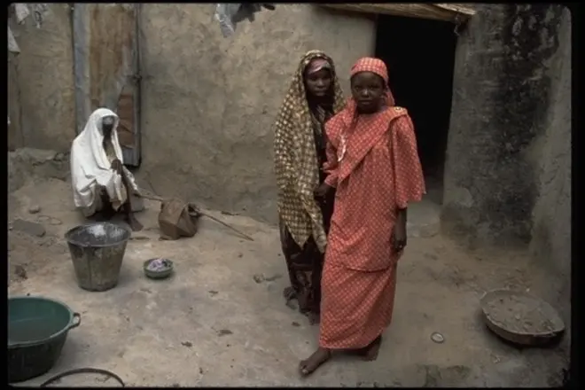 A young girl guiding an older woman by the hand past a seated wpam amd cooking equipment in an outdoor marketplace.
