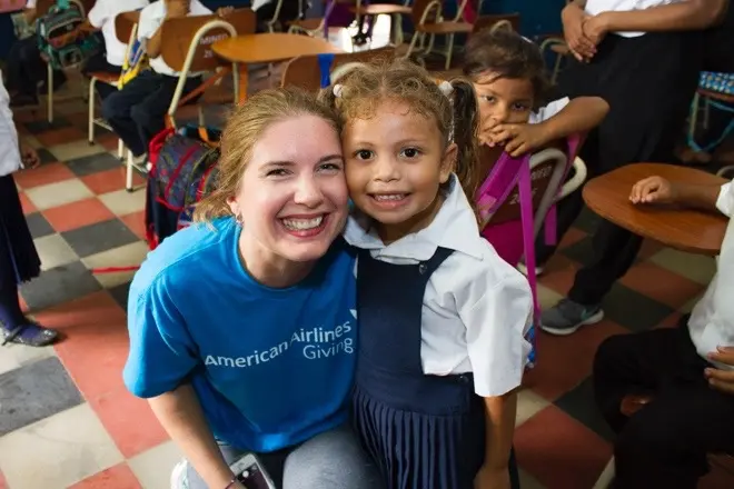 A smiling woman wearing a t-shirt that reads ‘American Airlines Giving’ kneels beside a smiling young girl who is wearing a school uniform in a classroom full of children.