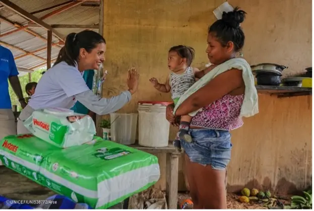 A smiling female aid worker holds her hand toward an infant who reaches a hand to her while being held by its mother. In the foreground, there are infant hygiene products on a table.