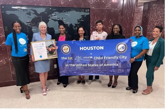 A group of adults and adolescents hold a plaque and banner, which reads 'Houston, the 1st Child Friendly City in the United States of America,’ at Houston City Hall.