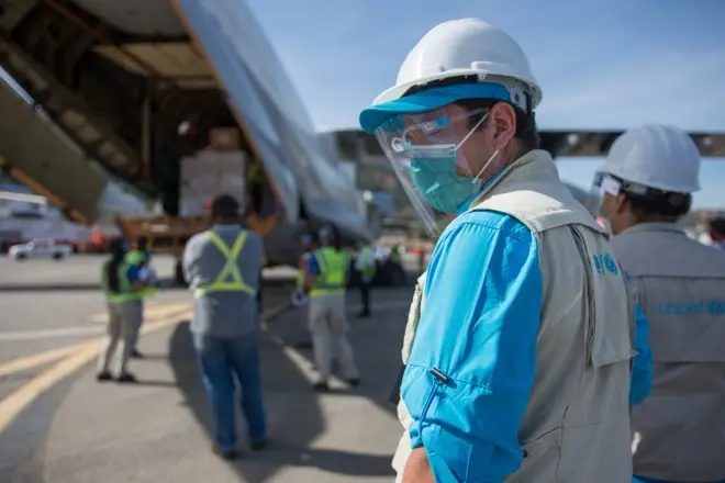 A man in UNICEF vest, helmet and plastic face shield standing near the open cargo hold of a plane, watching boxes being loaded into the hold.
