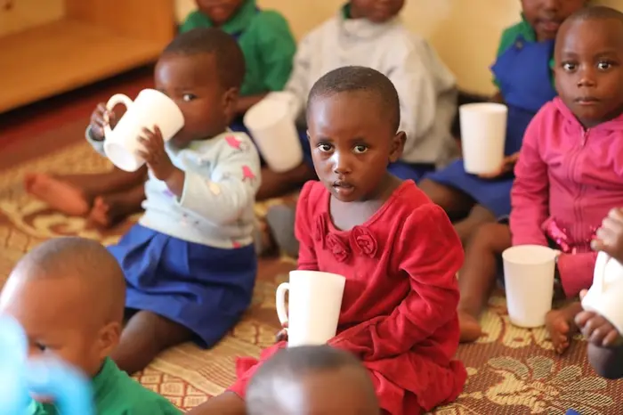 Young girl sitting on floor holding white mug looking directly into camera, surrounded by additional children holding and drinking from mugs