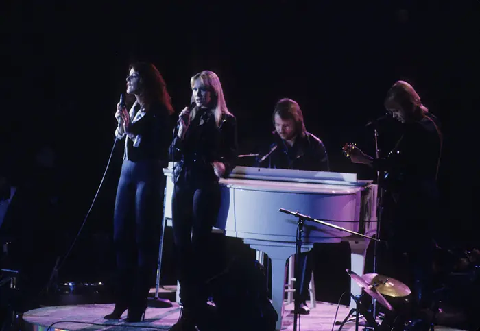 Abba’s four members dressed in black, singing into microphones on stage in front of a dark background.