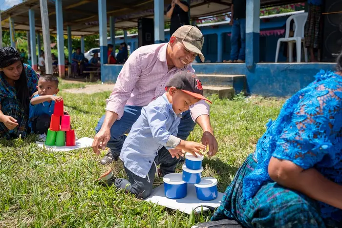 Young boy kneeling and stacking plastic cups while his father kneels behind him and assists