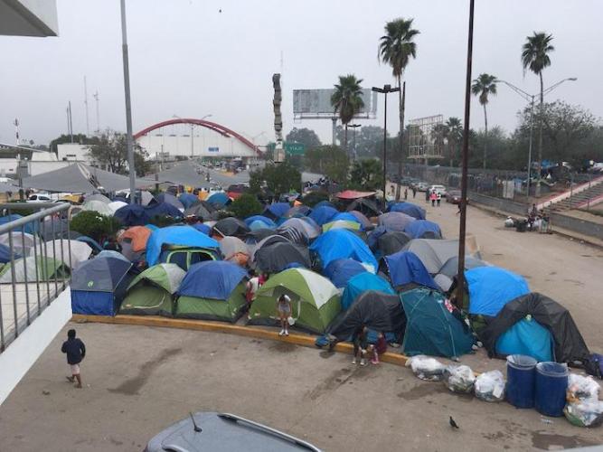 In December 2019, families seeking asylum in the U.S. live in a tent encampment in Matamoros, Mexico near the red-arched port of entry building, visible in the distance.