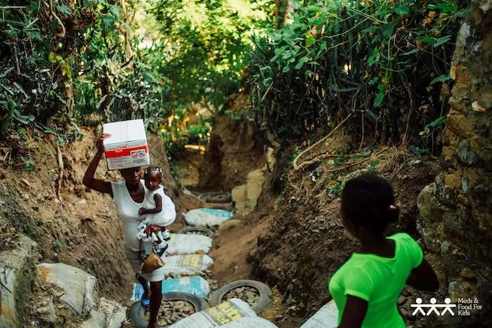 A mother carries her child and a carton of Ready-to-Use Therapeutic Food produced at the Meds & Foods For Kids factory in Haiti. A mother carries her child and a carton of Ready-to-Use Therapeutic Food produced at the Meds & Foods For Kids factory in Haiti.