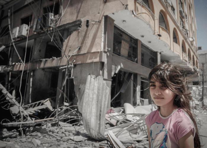 On May 17, 2021 in Gaza City, a Palestinian girl stands outside her family's damaged home while her parents hurry to remove some of their belongings. 