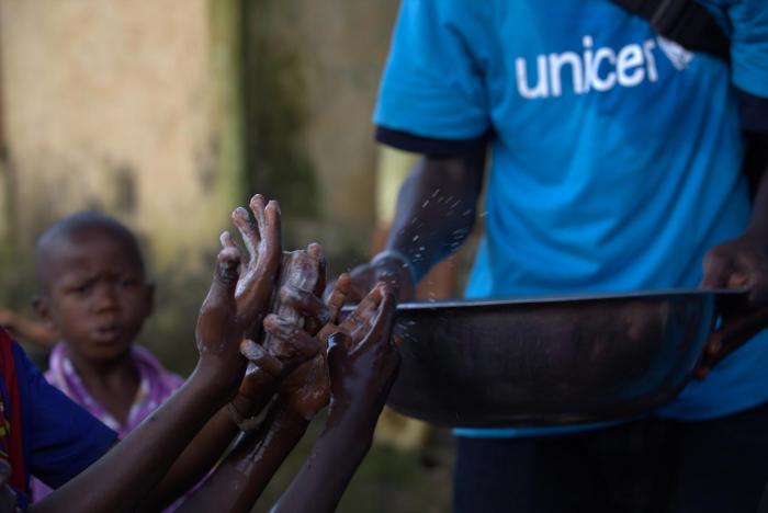 A social mobilizer from AJCOM, a UNICEF NGO partner, carries a basin of water past the soapy hands of children in Conakry, Guinea. © UNICEF/NYHQ2014-1522/La Rose
