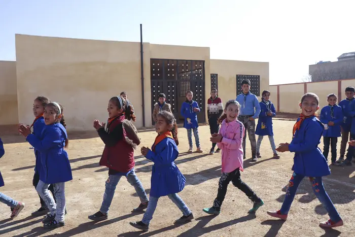 14 children smiling and playing outside in a sunny enclosed area. 