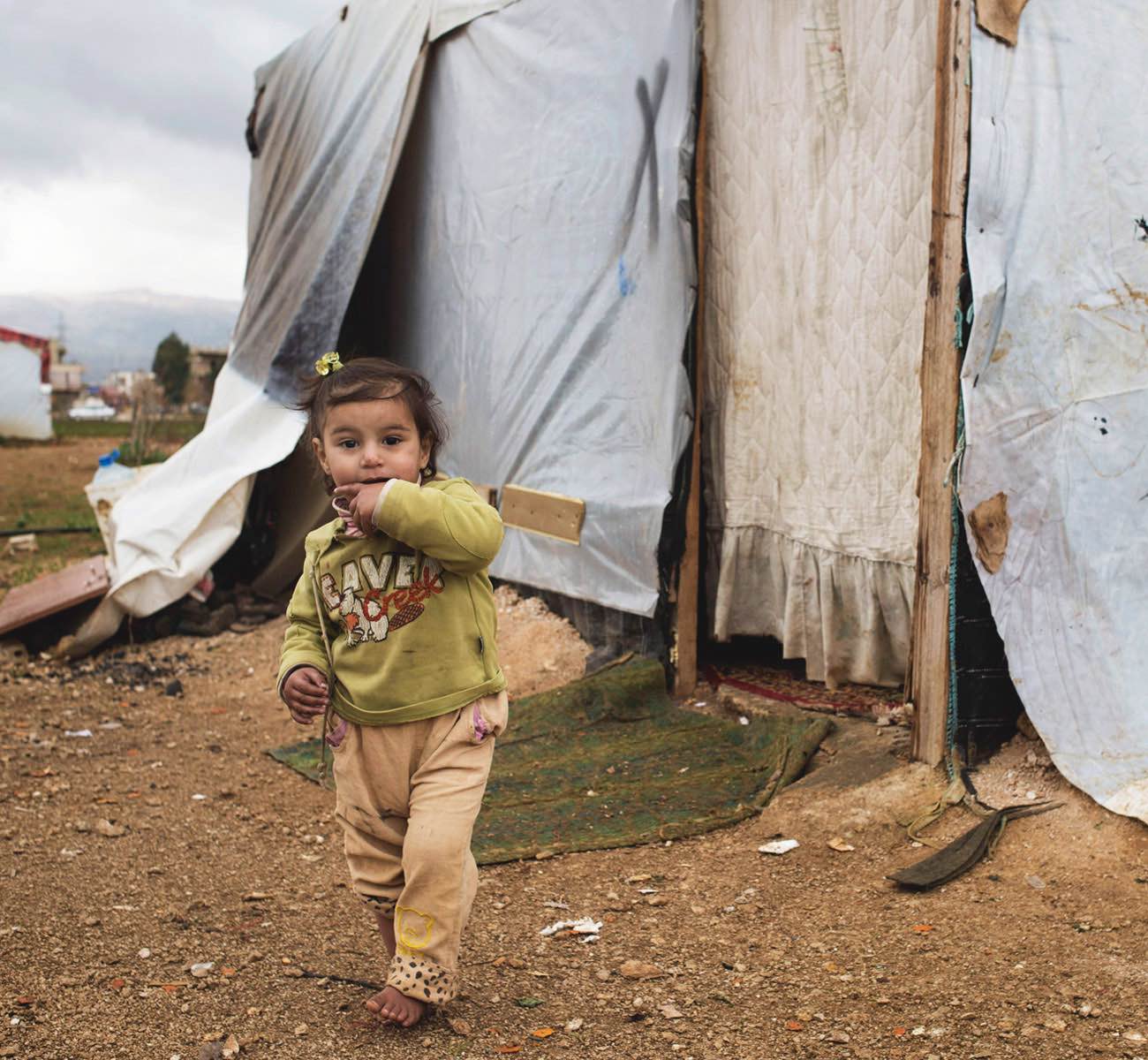 A young Syrian Refugee girl stands barefoot in the dirt outside of a rag-tag tent in a refugee camp in Jordan