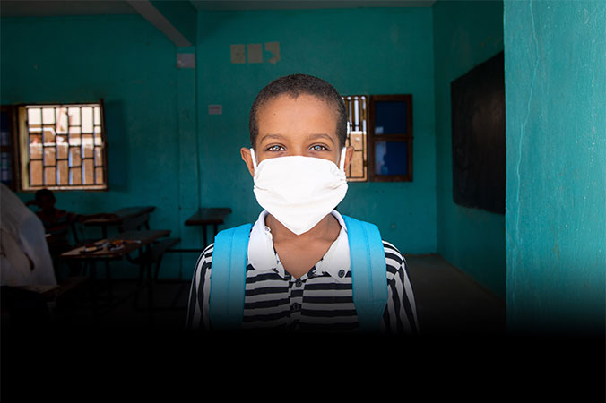 a boy in a school setting smiles while wearing a backpack and face mask